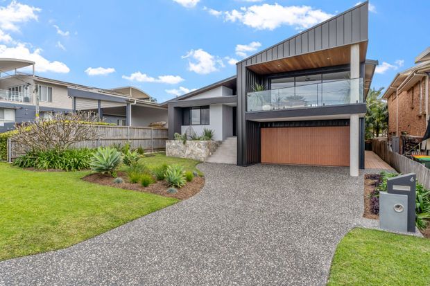 Contemporary multi-level residential house viewed from the street, featuring a dark grey rendered facade, vertical metal cladding, timber-lined upper balcony, expansive glazing, and a warm timber garage door. A broad exposed-aggregate driveway leads through a landscaped front garden with lawn and low-maintenance planting.