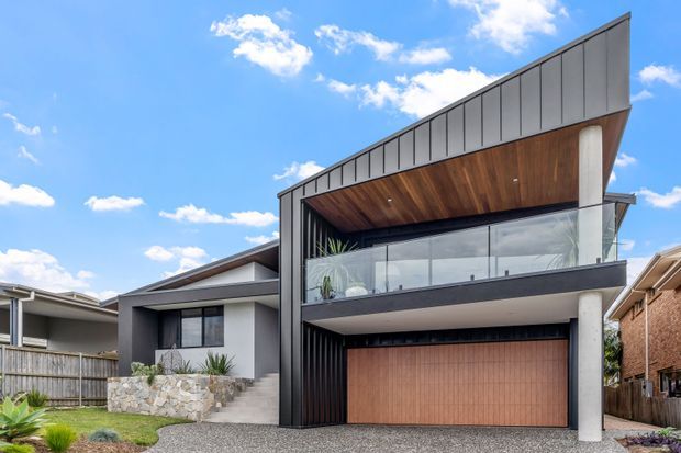 Contemporary two-storey residential facade with a dramatic angled roofline, dark vertical cladding, concrete columns, warm timber soffit, upper-level glass balcony, and a timber-toned double garage door. Stone landscaping and neighbouring suburban homes frame the exterior.