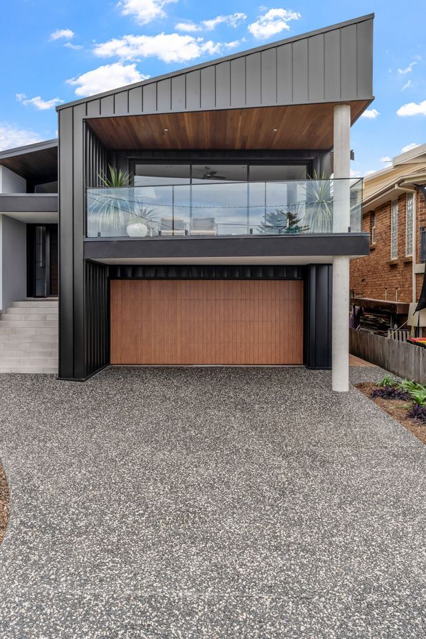 Street-facing exterior of a contemporary multi-storey residence with a dramatic angled parapet roofline, dark vertical metal cladding, a warm timber-lined upper balcony soffit, frameless glass balustrade, and a prominent white cylindrical support column. A timber garage door sits beneath the balcony, opening onto a textured aggregate driveway. Neighbouring homes are visible at the sides.
