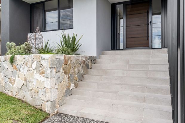 Front entry of a contemporary residential home, featuring broad pale stone steps leading to a timber pivot door with black-framed sidelights and glazing. The facade combines smooth light render with dark vertical cladding, while a rough natural-stone retaining wall and drought-tolerant planting define the entry landscape.