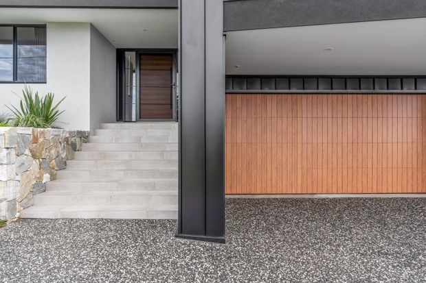 Modern residential exterior showing a recessed entry reached by broad pale tiled steps, a natural rough-stone retaining wall, dark rendered walls and structural column, and a warm timber-clad garage door beneath a concealed flat roofline. The foreground driveway has a speckled aggregate finish.
