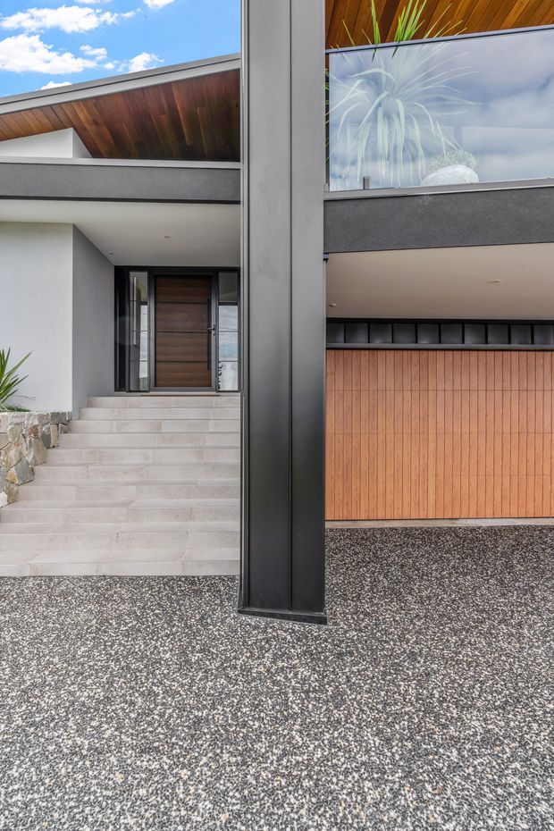 Exterior view of a contemporary residential entrance and garage frontage. The facade combines dark textured rendered bands, white walls, vertical timber garage cladding, a broad black structural column, pale tiled entry steps, and warm timber soffits. Glazing reflects the sky and reveals indoor planting, while the foreground is finished with a speckled aggregate surface.