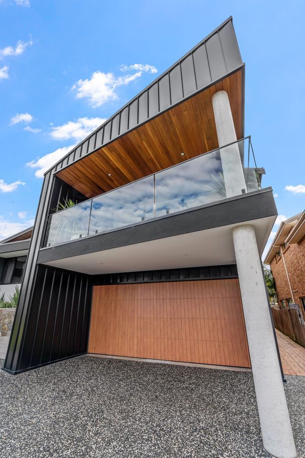 A contemporary multi-level residential facade viewed from the driveway, featuring a sharply pitched roofline, black vertical metal cladding, a warm timber-lined soffit, timber garage door, concrete support columns, and a glass-balustraded upper balcony.