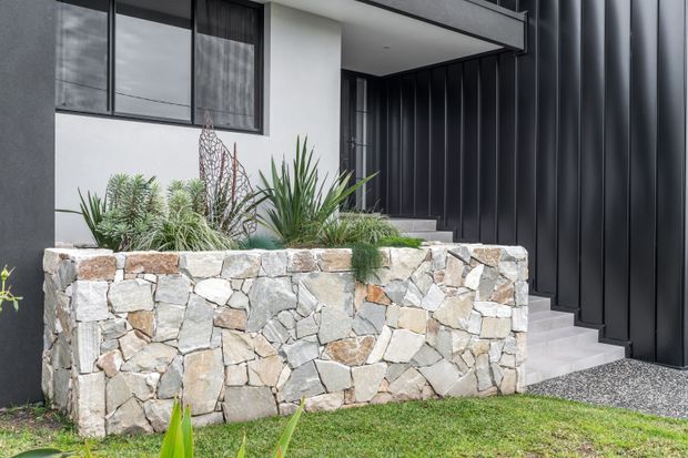 Contemporary residential entry facade featuring dark vertical metal cladding, a recessed covered porch, large black-framed glazing, pale concrete steps, and a raised planter retaining wall faced in irregular natural stone. Drought-tolerant ornamental planting softens the crisp monochrome exterior.