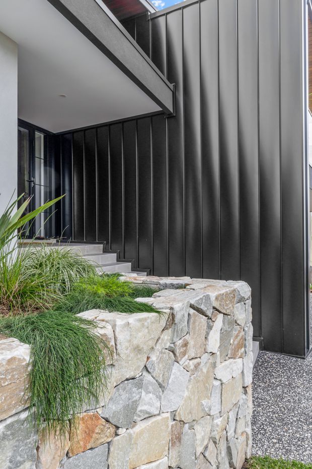 A contemporary residential exterior entry area featuring dark vertical metal cladding, a covered balcony or overhang, black-framed glazing, concrete steps, and a rough natural-stone retaining wall integrated with ornamental grasses and planting. The composition highlights the contrast between crisp dark facade panels and textured masonry landscaping.