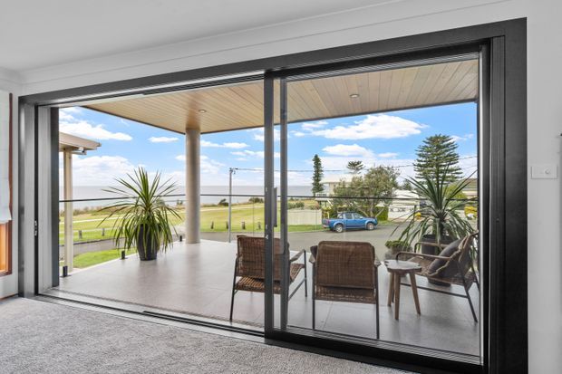 Interior view from a carpeted room through large black-framed sliding glass doors to a covered outdoor terrace. The terrace has a timber-lined ceiling, concrete columns, outdoor seating, and potted plants, with a bright coastal or waterfront landscape visible beyond.