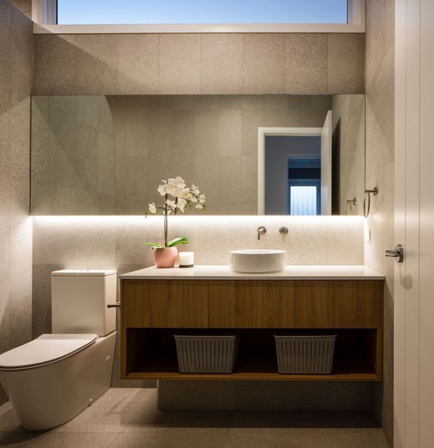 Contemporary bathroom featuring a wall-mounted timber vanity with open lower shelving, white vessel basin, wall-mounted tapware, integrated under-mirror lighting, large-format grey stone-look wall and floor tiles, and a wall-hung toilet. A large mirror and high-level window bring light into the compact space.