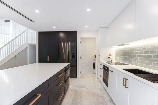 Bright contemporary galley kitchen with a large white island, contrasting dark lower cabinetry and tall black pantry/refrigerator cabinetry, white upper cabinets, integrated under-cabinet lighting, and a grey-green subway tile splashback. A staircase and laundry area are visible beyond the kitchen.