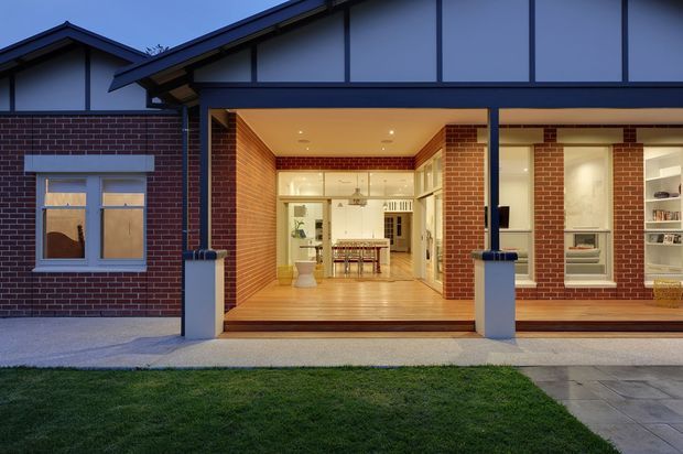 Front exterior view of a single-storey brick home at dusk, with a large open glazed entry or living opening framed by dark timber or metal beams. The illuminated interior reveals a timber-floored open-plan kitchen and dining area, while the facade combines red brick, pale rendered panels, and exposed structural framing.