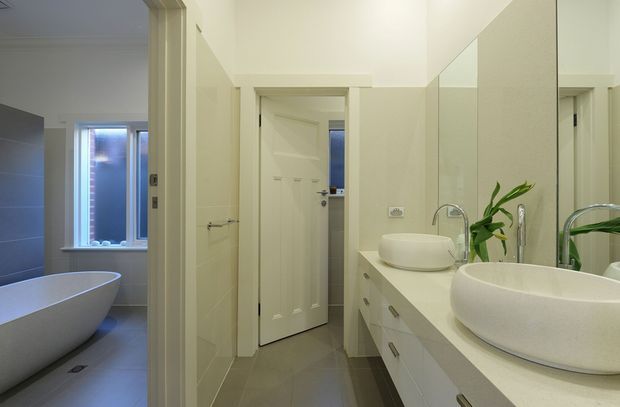 A bright contemporary bathroom vanity area with two vessel basins, pale stone benchtop, white drawers, wall-to-wall mirror, and chrome fixtures. An open doorway reveals a separate bathtub area with a freestanding white bath and frosted window. The room uses a restrained palette of white, cream, light grey, and chrome, with recessed ceiling lighting and minimal styling.