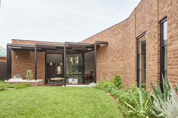 Contemporary single-storey brick residence viewed from a landscaped courtyard or garden. The facade features warm brown exposed brick, tall dark-framed windows, and a glazed central entry or living connection beneath a slim steel pergola with a slatted roof. A paved outdoor sitting area and low planted garden border the building, creating a sheltered courtyard setting.