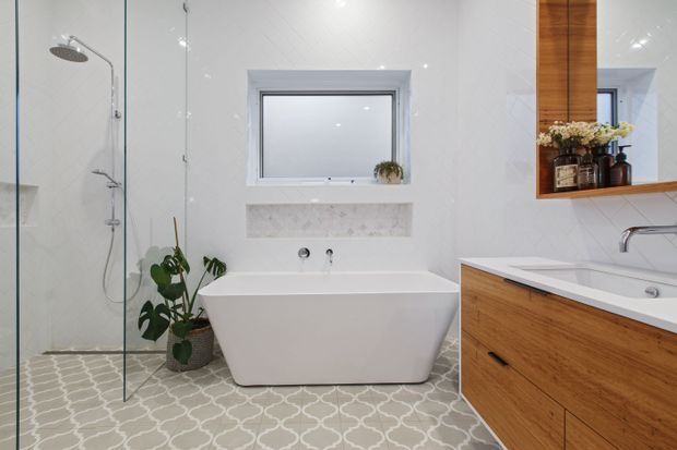 Bright contemporary bathroom featuring a freestanding white bathtub beneath a frosted window, a glass-enclosed walk-in shower, patterned grey-and-white floor tiles, and warm timber vanity cabinetry with a matching mirrored storage unit. White tiled walls and recessed shelving create a clean, refined appearance, with a small plant adding an organic accent.