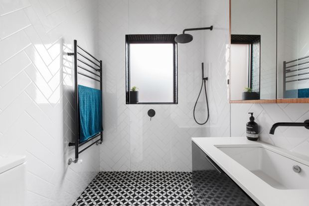 A contemporary bathroom with white geometric wall tiles, a black-and-white patterned floor, black-framed window, black shower fittings, a wall-mounted towel warmer holding a blue towel, and a white freestanding bathtub beneath a large mirrored vanity.