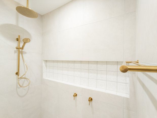 Bright contemporary bathroom shower enclosure with large pale wall tiles, a recessed tiled niche, and brushed gold shower fittings, including an overhead rain shower, hand shower, controls, and towel rail.