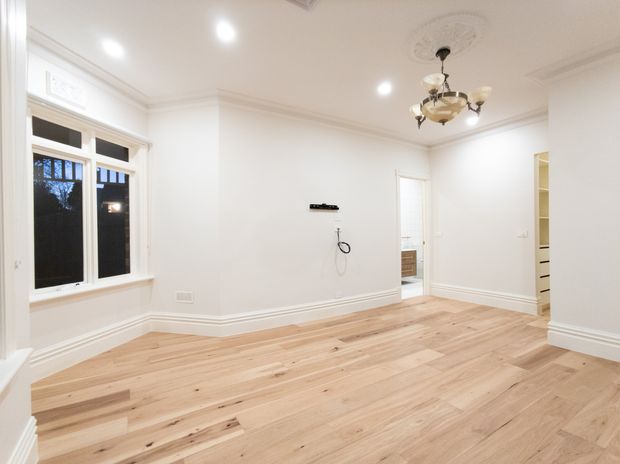 Bright, unfurnished traditional-style room with wide-plank light timber flooring, white walls, decorative ceiling cornice and ceiling medallion, recessed downlights, and a classic chandelier. A large framed window and open doorway provide natural light, while built-in shelving or cabinetry is visible at the right.