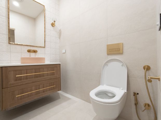 A compact contemporary bathroom with a wall-hung timber vanity, integrated pale countertop basin, large gold-framed mirror, light beige tiled walls and floor, and a white wall-mounted toilet with brass fixtures. The palette is warm and neutral with refined metallic detailing.