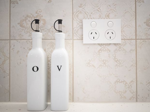 Close-up of two white pump bottles labelled “O” and “V” standing on a pale countertop against a decorative cream floral tiled wall, with a white double electrical outlet visible to the right.