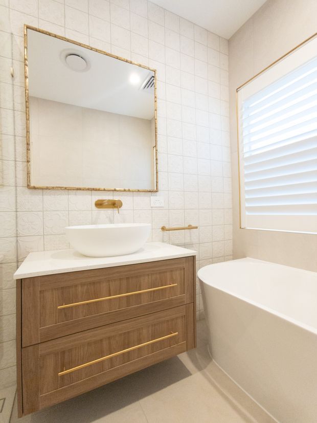 Bright contemporary bathroom featuring a floating timber vanity with long brass drawer pulls, a white vessel basin, white tiled walls with a subtle decorative patterned band, a gold-framed mirror, wall-mounted gold tapware, and a freestanding white bathtub beside a shuttered window.