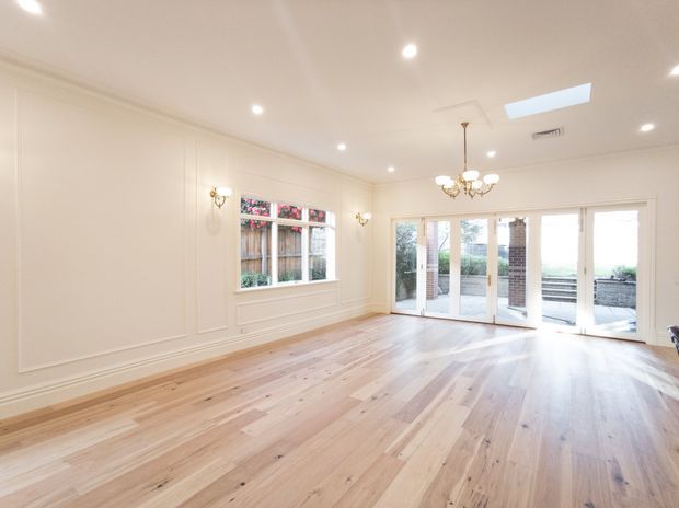 Bright, mostly empty formal living or dining space with pale timber flooring, white decorative wall panelling, French doors opening to an outdoor terrace, a large window, chandeliers, wall sconces, recessed lighting, and a skylight. The room has refined traditional detailing and abundant natural light.