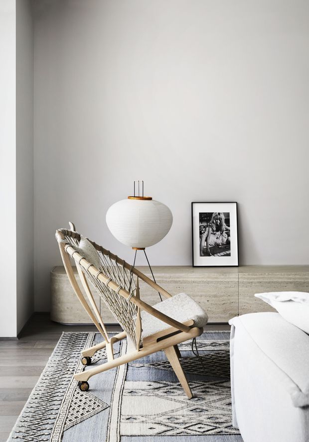 A softly lit contemporary living room corner featuring a woven timber lounge chair in the foreground, a low textured stone or concrete sideboard, a sculptural paper pendant light, framed black-and-white artwork, and a patterned area rug beside a white sofa. The palette is neutral with light timber, warm grey, white, and black accents.