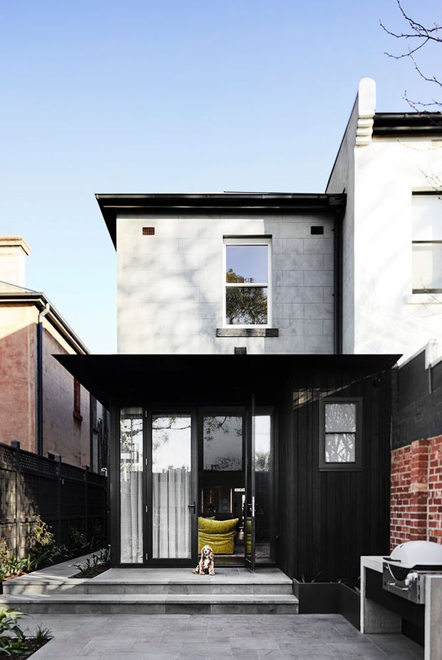 Exterior view of a narrow, multi-storey urban townhouse or terrace-house extension. The facade combines a white rendered upper level with a dark vertical-clad lower entry volume, broad black metal roof overhangs, large glazed entry doors, and a paved front terrace with steps. Adjacent brick walls and neighbouring buildings frame the compact site.