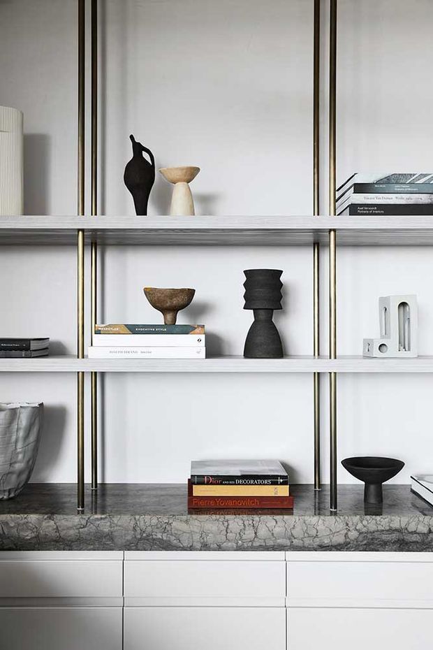 A refined built-in display and storage unit with white shelving, slim brass-toned vertical supports, and a dark veined stone benchtop over white drawers. The shelves are styled with books and sculptural ceramic and dark decorative objects, creating a minimalist, gallery-like interior vignette.