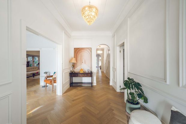 Bright, elegant residential hallway with white paneled walls, decorative ceiling mouldings, warm herringbone timber flooring, and a central console beneath a framed artwork. A sculptural pendant light hangs overhead, while open doorways reveal adjoining furnished rooms and an arched passage extends down the corridor. A potted plant and upholstered chair add soft furnishings in the foreground.