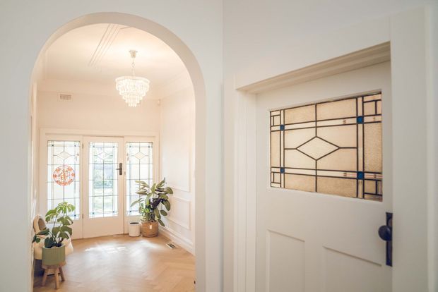 Bright traditional entryway featuring a white paneled front door with decorative stained-glass sidelighting, opening toward a small vestibule with an arched opening, chandelier, leadlight glass doors, potted plants, and a warm herringbone timber floor.