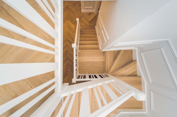 A top-down view into a bright, multi-level residential stairway. White painted balustrades and moulded wall panels frame timber stairs, including a contrasting herringbone-patterned landing and warm pale timber treads. The composition emphasizes the stairwell's geometric railings and layered flights.