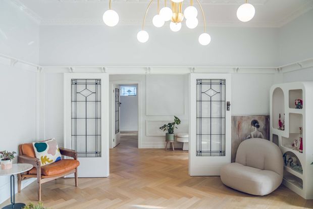 Bright, refined living room with white walls and ornate ceiling mouldings, parquet herringbone flooring, glazed geometric-panel double doors, built-in arched display shelving, and a sculptural pendant light. The room is furnished with a curved lounge chair, mid-century-style armchair, and small side table, creating a playful yet elegant interior.