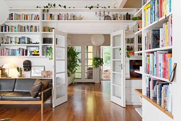 Bright, book-lined interior looking through open glazed double doors into a central hallway or entry passage. White built-in bookshelves span both sides and contain books, plants, and decorative objects. A compact living area with a black leather sofa and side table is visible on the left, while polished medium-toned timber flooring continues through the space. The room has white painted joinery and trim, abundant daylight, indoor greenery, and a warm, lived-in library-like atmosphere.