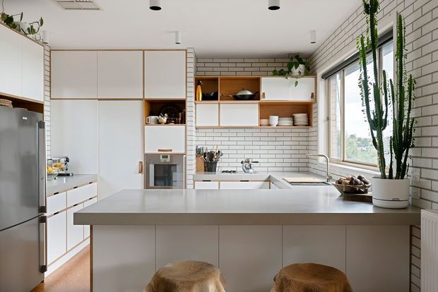 Bright contemporary kitchen with a large central island and two stools, white cabinetry, open timber shelving, white brick-style wall tiles, stainless-steel appliances, and large windows. Tall indoor plants add greenery beside the glazing, while recessed and pendant lighting create a clean, airy atmosphere.
