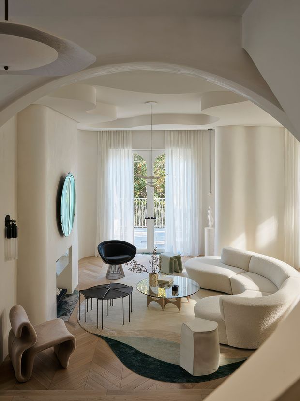 A refined, light-filled living room with sculptural curved architectural detailing, including an undulating ceiling and broad arched opening. The room features a curved cream sofa, sculptural lounge chairs, glass coffee table, pale herringbone timber flooring, sheer curtains, and a balcony or garden view through tall glazed doors. A rounded wall mirror, fireplace or recessed display niche, and soft neutral furnishings reinforce the elegant contemporary character.