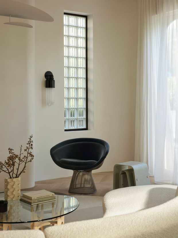 A refined, light-filled living room with sculptural contemporary furniture. The space features a curved black lounge chair, a cream sofa in the foreground, a glass coffee table, sheer full-height curtains, a tall narrow glass-block window with dark framing, pale walls, and herringbone timber flooring. Decorative branches and books add understated styling, while sculptural ceiling and wall lighting contribute to the minimalist, gallery-like atmosphere.