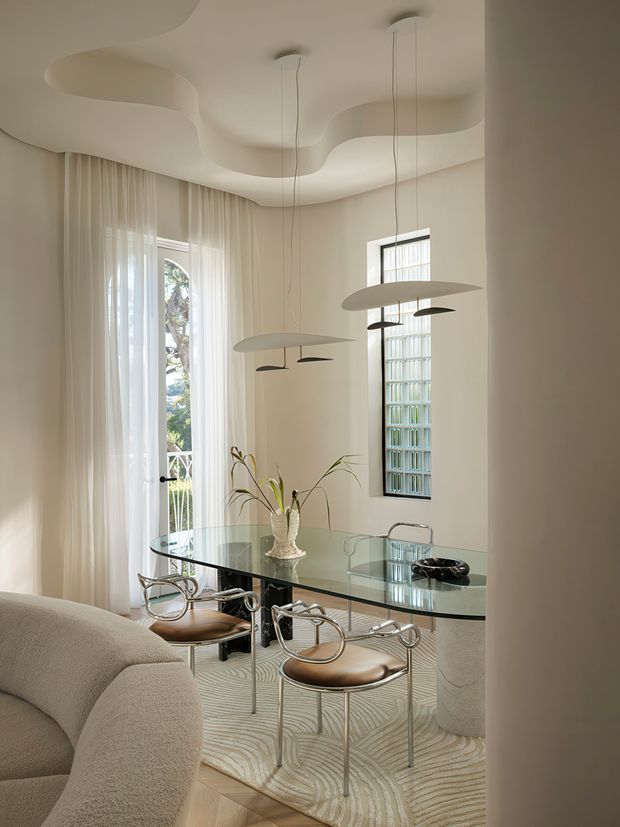 A refined dining area viewed from an adjacent space, featuring a glass dining table with sculptural metal-framed chairs, a curved upholstered sofa in the foreground, sheer floor-to-ceiling curtains, a textured wavy ceiling, a glass-block window, sculptural pendant lights, and a neutral cream palette.