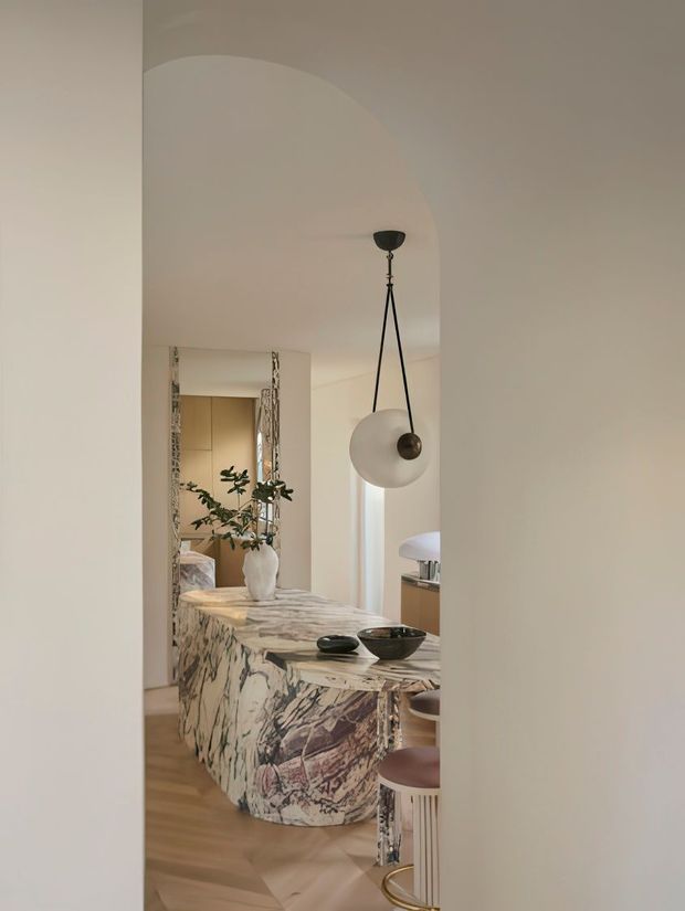 View through a wide interior opening into a refined kitchen and dining area. A sculptural marble-clad island with dramatic veining dominates the space, paired with rounded stools and a suspended black-and-white pendant light. A large decorative mirror reflects cabinetry, a plant, and part of the adjacent room. Warm timber flooring, soft off-white walls, and restrained contemporary furnishings create an elegant, calm interior.