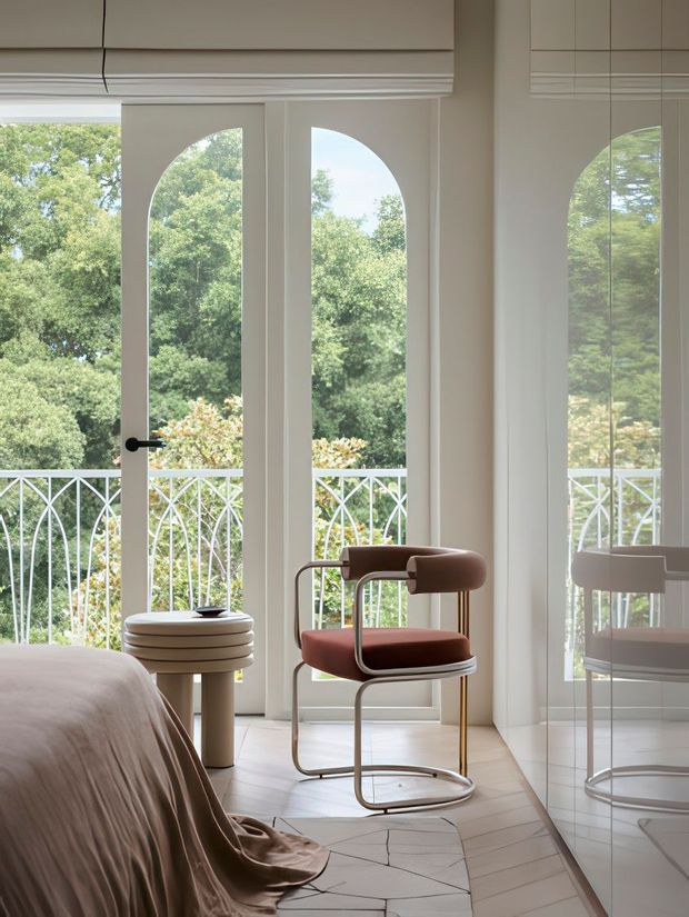 Bright, refined bedroom corner with full-height white-framed glazed French doors opening to a balcony with decorative white metal balustrades and leafy views. A sculptural rust-coloured upholstered chair sits beside a small round stacked side table, while a bed with soft neutral bedding occupies the foreground. A mirrored wall reflects the chair and glazing. The room has pale herringbone flooring, white walls, and layered Roman blinds.