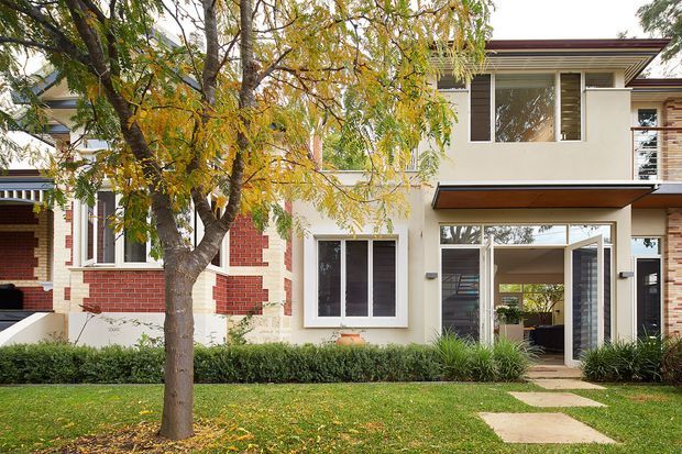 Street-facing view of a contemporary two-storey residential house with a mixed brick and smooth rendered facade, broad glazed openings, a projecting upper-level canopy, and an open ground-floor entry or living area. The home sits behind a neatly landscaped front lawn with a mature deciduous tree, low hedging, ornamental grasses, and stepping-stone paving.