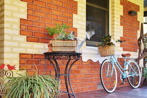 A colourful residential porch featuring red brick walls with cream mortar and contrasting cream brick bands, a dark-framed window, decorative wrought-iron plant stand, potted plants, and a vintage light-blue bicycle with a wicker basket. The space has a warm, nostalgic character and timber decking.