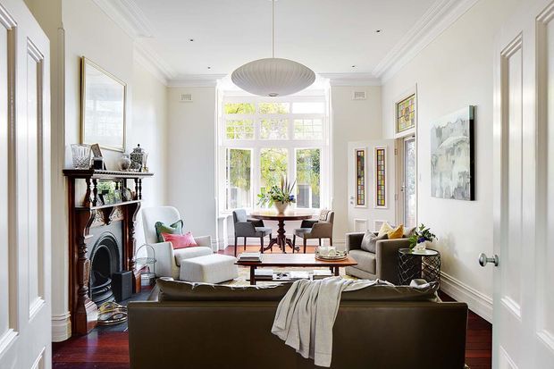 Bright, elegant living room in a refined traditional interior, viewed through open double doors. The room features high ceilings with decorative crown moulding, white paneled walls and doors, dark timber flooring, a classic fireplace with mantel, a large central pendant light, and a seating arrangement oriented around a coffee table. Tall multi-pane windows bring in abundant natural light, with a small dining area positioned near the rear window.