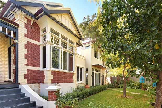 A sunny view of a multi-storey residential house with a prominent gabled front elevation, red and cream brickwork, cream painted timber detailing, dark trim, projecting bay windows, and a small upper balcony. A later white-rendered extension with tall glazing sits behind the original facade, opening toward a landscaped lawn and mature trees.