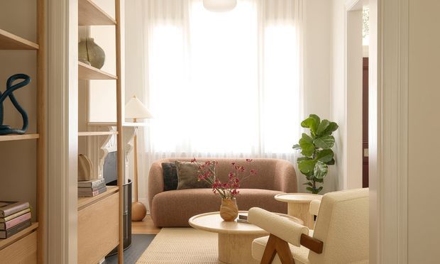 A warm, softly lit living room with a curved caramel-brown sofa positioned in front of tall sheer-curtained windows. Built-in timber shelving and cabinetry line the left side, while a rounded cream coffee table, textured rug, sculptural cream armchair, indoor plant, and understated decorative objects create a refined, organic interior.