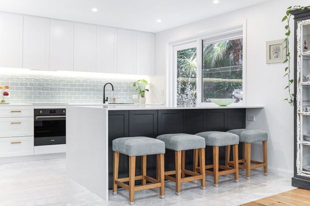 Bright contemporary kitchen with white upper and lower cabinetry, a pale green-grey subway tile splashback, and a dark island with a white benchtop. Four timber-framed stools with grey upholstered seats line the island, while a large window brings in views of greenery. The room has light flooring, recessed ceiling lights, and a clean, airy presentation.