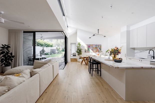 Bright, open-plan contemporary interior combining a living room and kitchen. The space features pale timber flooring, white walls and cabinetry, a large white island with integrated seating, expansive sliding glass doors opening to a landscaped outdoor area, a neutral sectional sofa, indoor plants, and colourful artwork and floral arrangements. A slim black linear pendant adds contrast above the island.