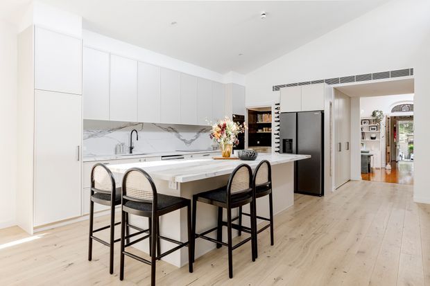 A bright, open-plan contemporary kitchen with extensive white flat-panel cabinetry, a large marble-look island with seating for four, integrated appliances, and pale timber flooring. Black-framed bar stools and dark hardware provide contrast. The kitchen opens toward a hallway and adjoining rooms, with a decorative vase of flowers on the island.