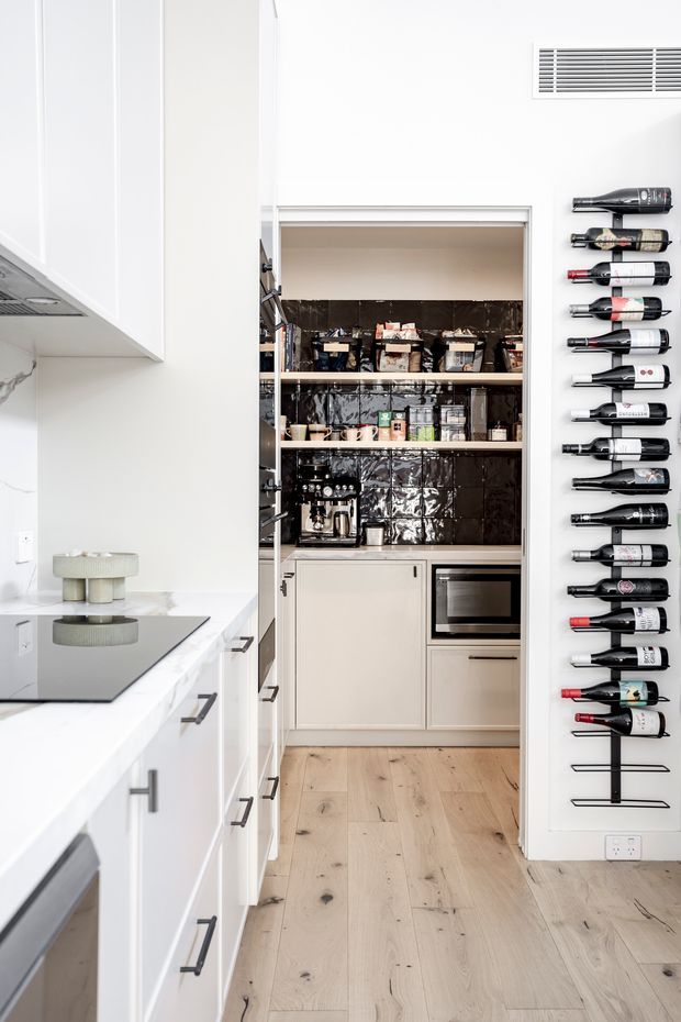 A bright, narrow contemporary kitchen focused on a built-in walk-in pantry. White shaker-style cabinetry and a white stone benchtop frame a black induction cooktop in the foreground. The pantry has dark glossy tiled walls, open shelving stocked with food and small appliances, integrated cabinetry, and a built-in stainless-steel oven. A wall-mounted wine rack displays numerous bottles beside the pantry, while pale timber flooring runs throughout.