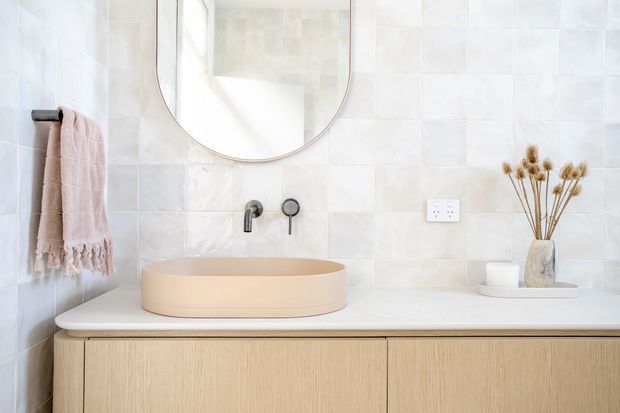 Bright contemporary bathroom vanity with a pale beige vessel basin on a white countertop, light timber floating cabinetry, glossy white square wall tiles, brushed dark metal wall-mounted tapware, a rounded frameless mirror, and a soft neutral towel with dried botanical decor. The composition is clean, warm, and minimalist.