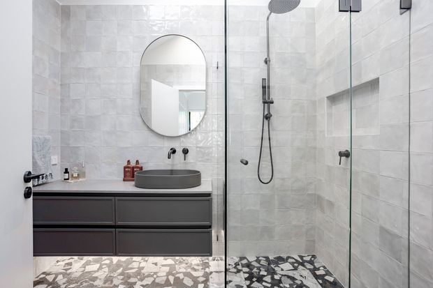 A contemporary bathroom with a dark floating vanity, round vessel basin, wall-mounted black tapware, arched round-edged mirror, and a frameless glass shower enclosure. The walls and shower are clad in pale textured rectangular tiles, while the floor features a bold monochrome irregular mosaic pattern.
