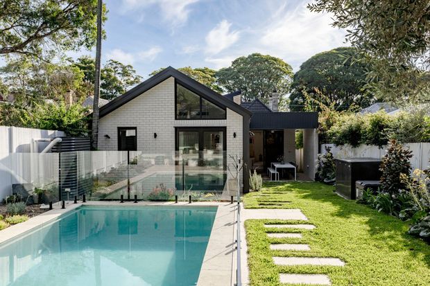 Exterior view of a contemporary residential home with a prominent steep gabled volume clad in light brick, large glazed openings, and a darker flat-roofed extension. A swimming pool with frameless glass fencing occupies the foreground, alongside a lawn and stepping-stone path. Mature landscaping surrounds the property.
