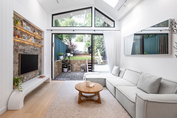 Bright contemporary living room with a large light-grey sectional sofa, round timber coffee table, pale timber flooring and a textured neutral rug. A white curved TV console and exposed brick fireplace wall with built-in timber shelving form the main feature. Black-framed floor-to-ceiling glazing opens to a compact landscaped courtyard, while a high angled ceiling and clerestory windows bring in abundant daylight.